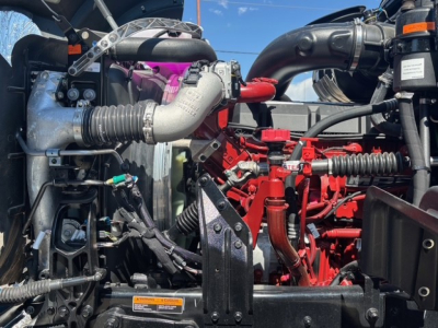 Engine bay of a 2026 Mack GR64F dump truck showing turbocharger, intake piping, red engine components, hoses, and various labels.