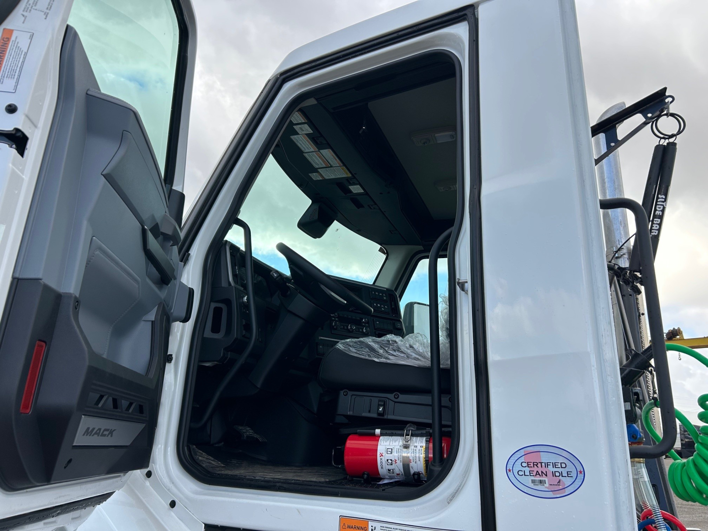 Cab interior of a new 2026 Mack PR64T daycab, with open door, steering wheel, dashboard, and a red fire extinguisher visible on the floor.