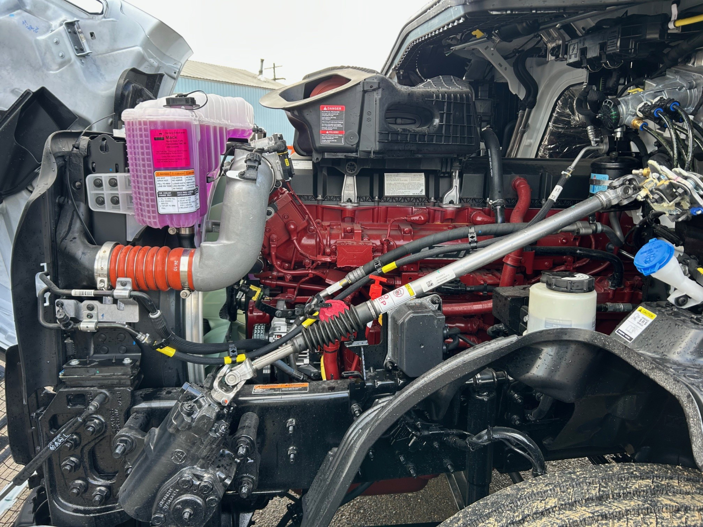 Mack PR64T engine bay showing red engine block, pink coolant reservoir, hoses, and various components.
