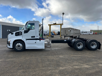 Truck chassis side view showing white 2026 Mack PR64T daycab with long wheelbase and dual rear axles, empty flatbed rail, and chrome exhaust stack.