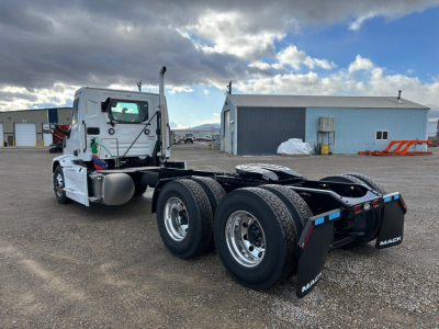 Mack PR64T truck chassis with dual rear axles and white cab, visible fuel tanks, exhaust stack, and chrome wheels; exterior setting with industrial buildings in background.