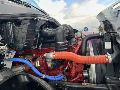 Mack PR64T engine bay showing red engine block with orange flexible hose, blue hose, black plastic intake, and surrounding components.