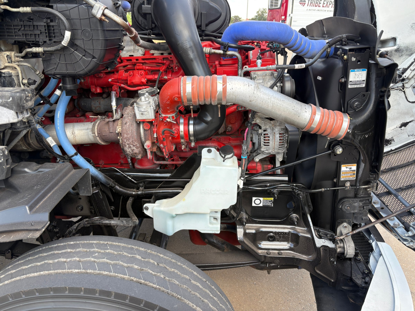 Peterbilt 579 engine bay with red engine block, blue hoses, silver exhaust, and white coolant reservoir visible from side view.