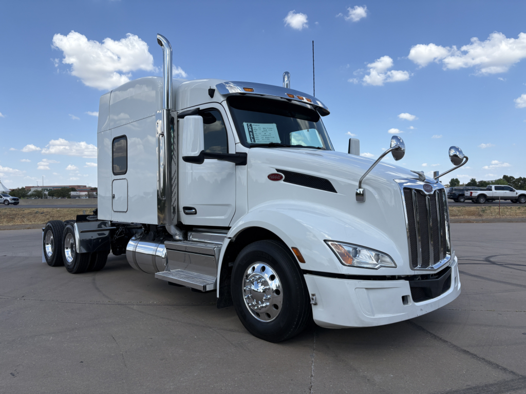 Peterbilt 579 tractor truck in cab-forward configuration, white exterior, sleeper, dual rear axles, large chrome exhaust stack, chrome mirrors, daytime running lights, front grille with Peterbilt badge.