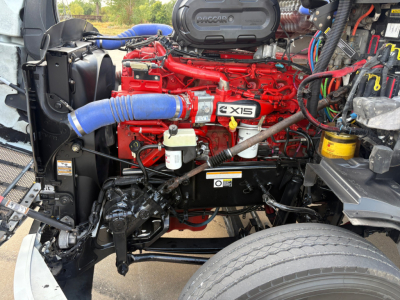 Peterbilt 579 engine bay with red engine, blue hoses, and black chassis. Visible components include radiator, intercooler piping, air intake, filters, wiring harnesses, and front wheel assembly.