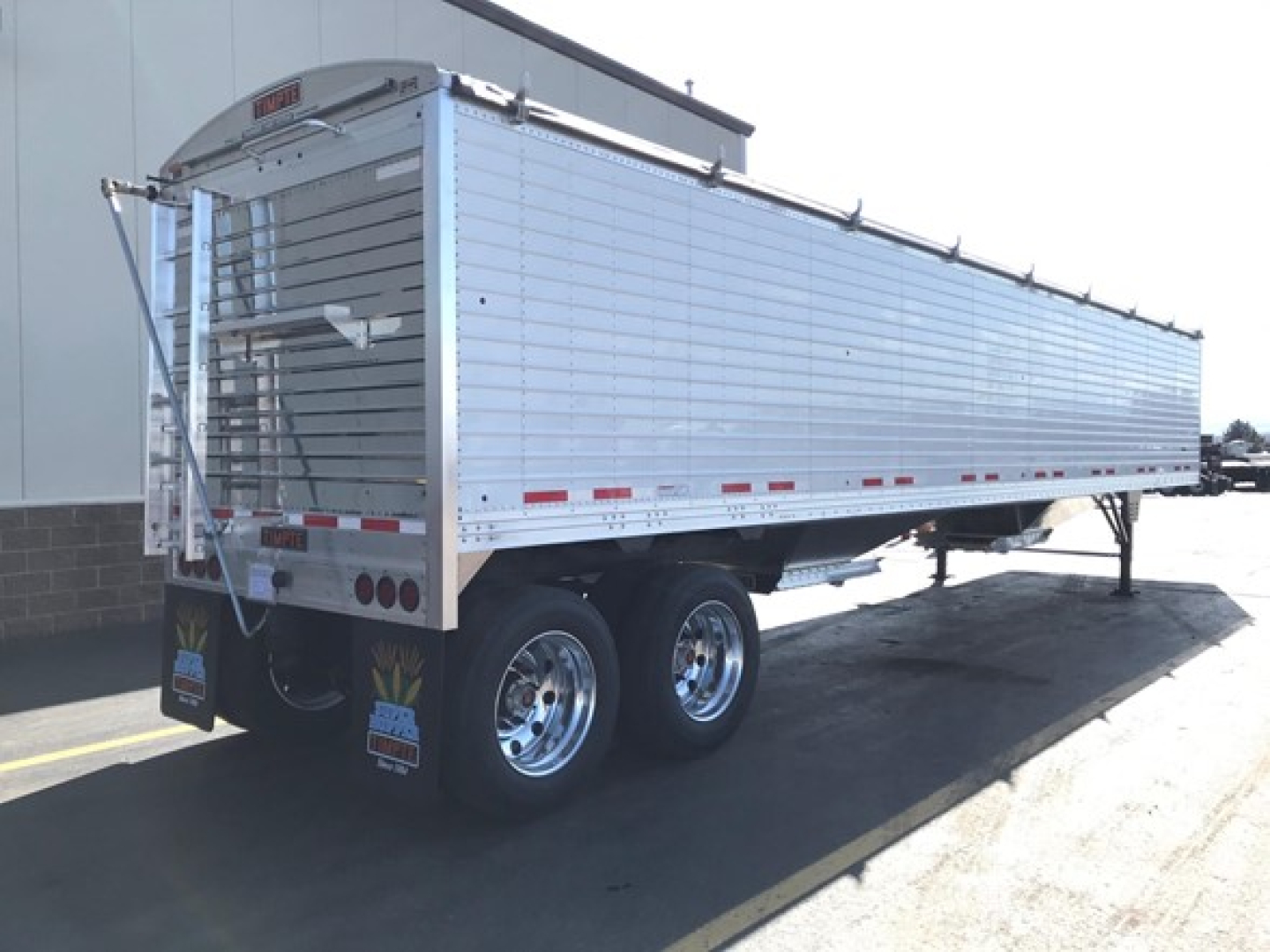 Trailer side and rear showing Timpte Super Hopper trailer with dual rear wheels, aluminum-sided body, and rear gate. Visible text includes Timpte branding on rear license panel and reflective striping.