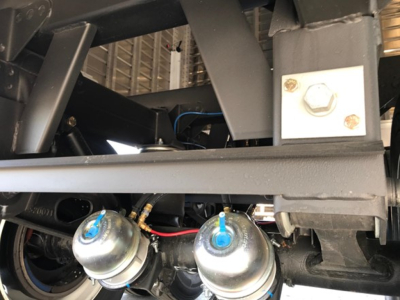 Trailer component close-up showing air clamps and hydraulic lines in a Timpte Super Hopper trailer chassis. Visible hardware includes blue-capped fittings, metal brackets, and a bolt.