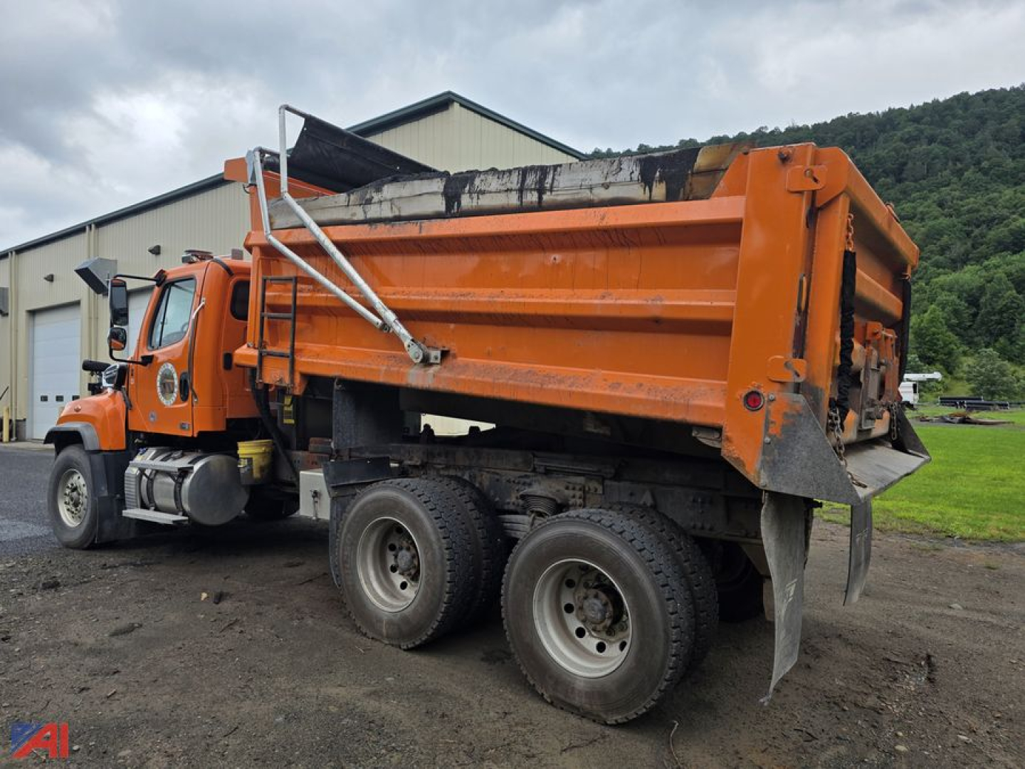 Dump truck 2020 Freightliner 114 SD with orange dump bed viewed from rear-left side, dual rear axles, black undercarriage, sliding tailgate, in yard.