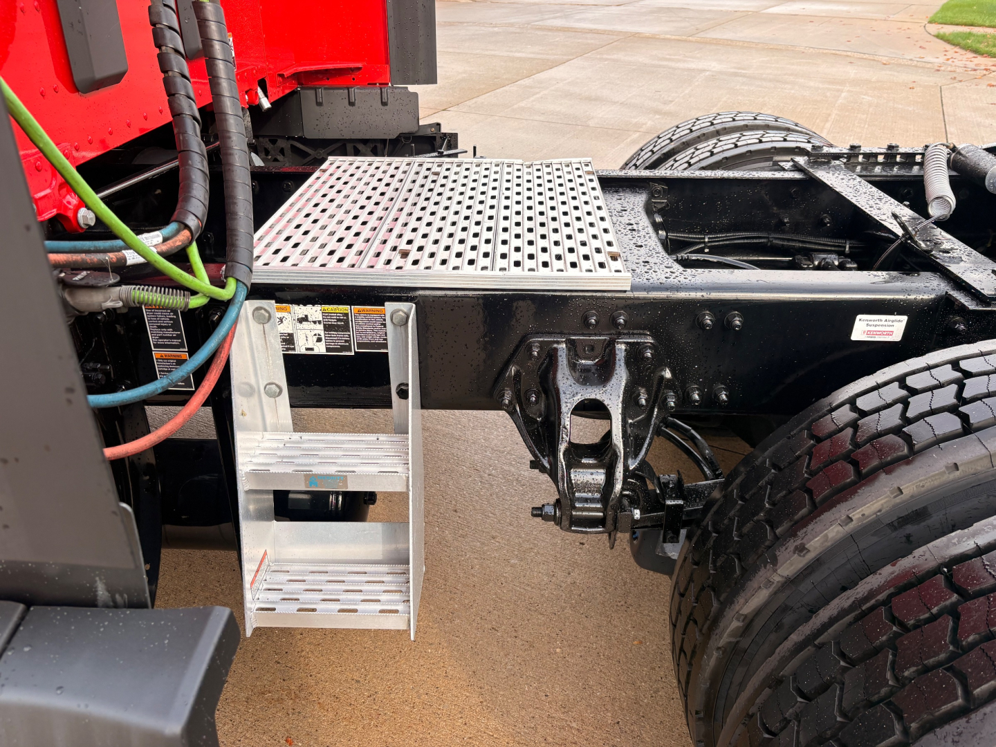 Kenworth T680 chassis area showing aluminum step, hydraulic lines, and rear dual tires with drivetrain components visible.