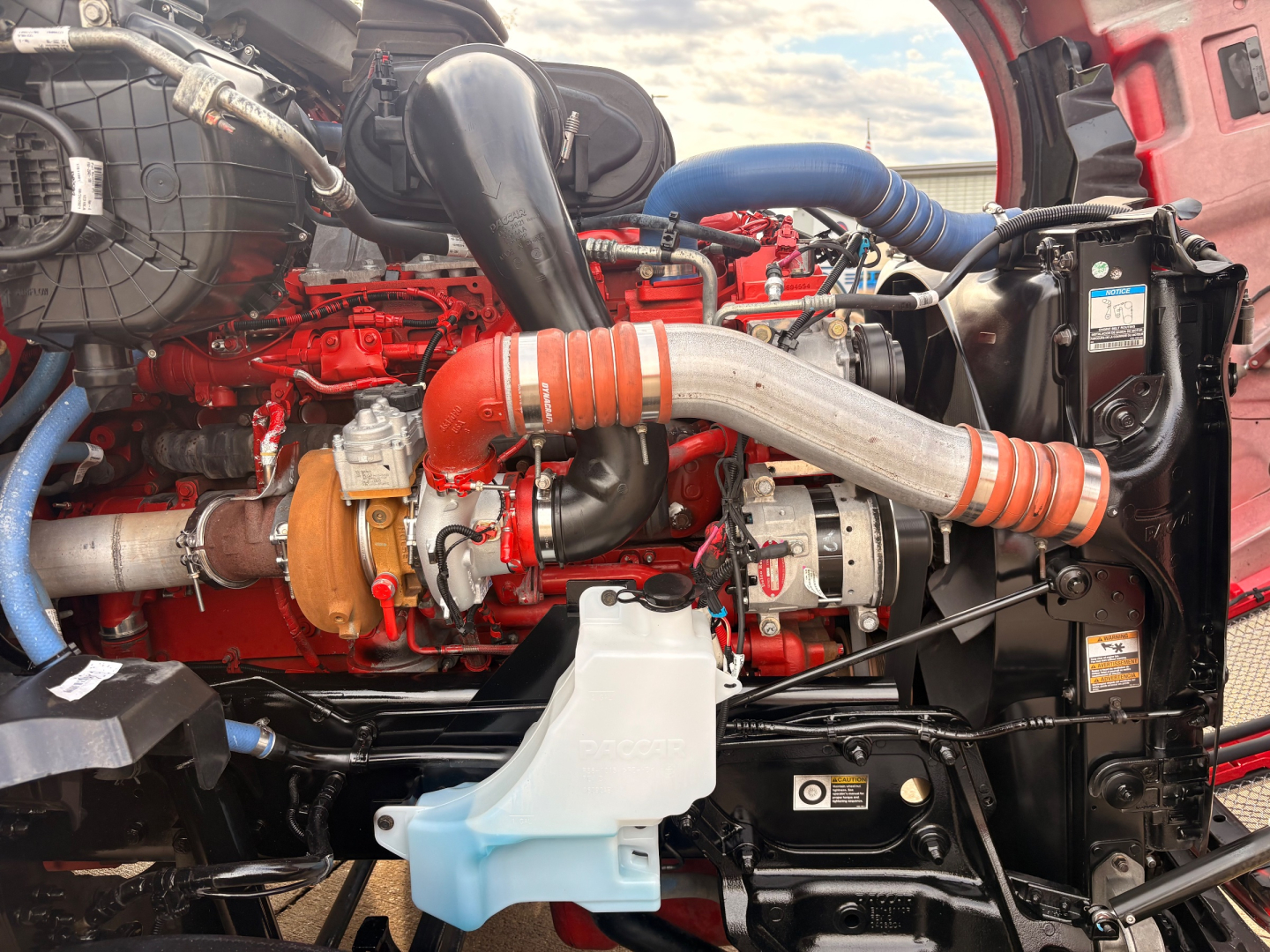 Engine bay of a 2022 Kenworth T680 with red engine, blue hoses, orange intake piping, and assorted labels visible.