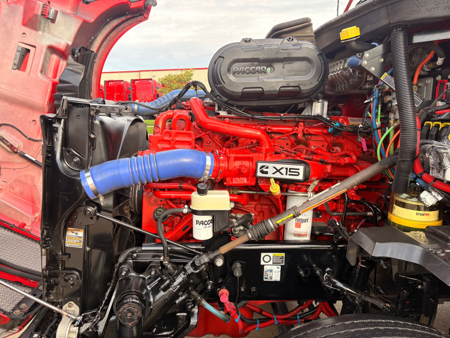 Kenworth T680 engine bay showing Cummins X15 engine with blue intercooler hose, white reservoir, and various red components.