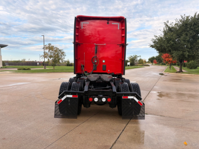 Rear view of a red Kenworth truck cab with black mud flaps and dual rear wheels, showing electrical connectors and hitch area.