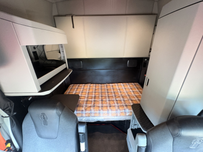 Kenworth T680 truck sleeper cab interior showing bed platform with plaid mattress, storage cabinets, and seating in foreground.