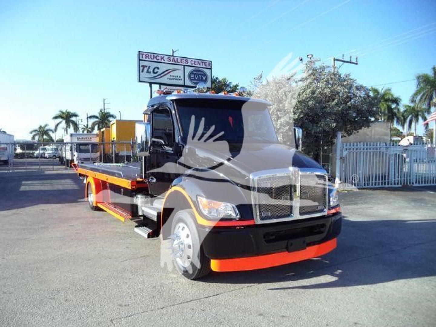 Kenworth T280 flatbed rollback truck in a dealership lot, front view with black cab and orange accents, stock number and dealership sign in background.