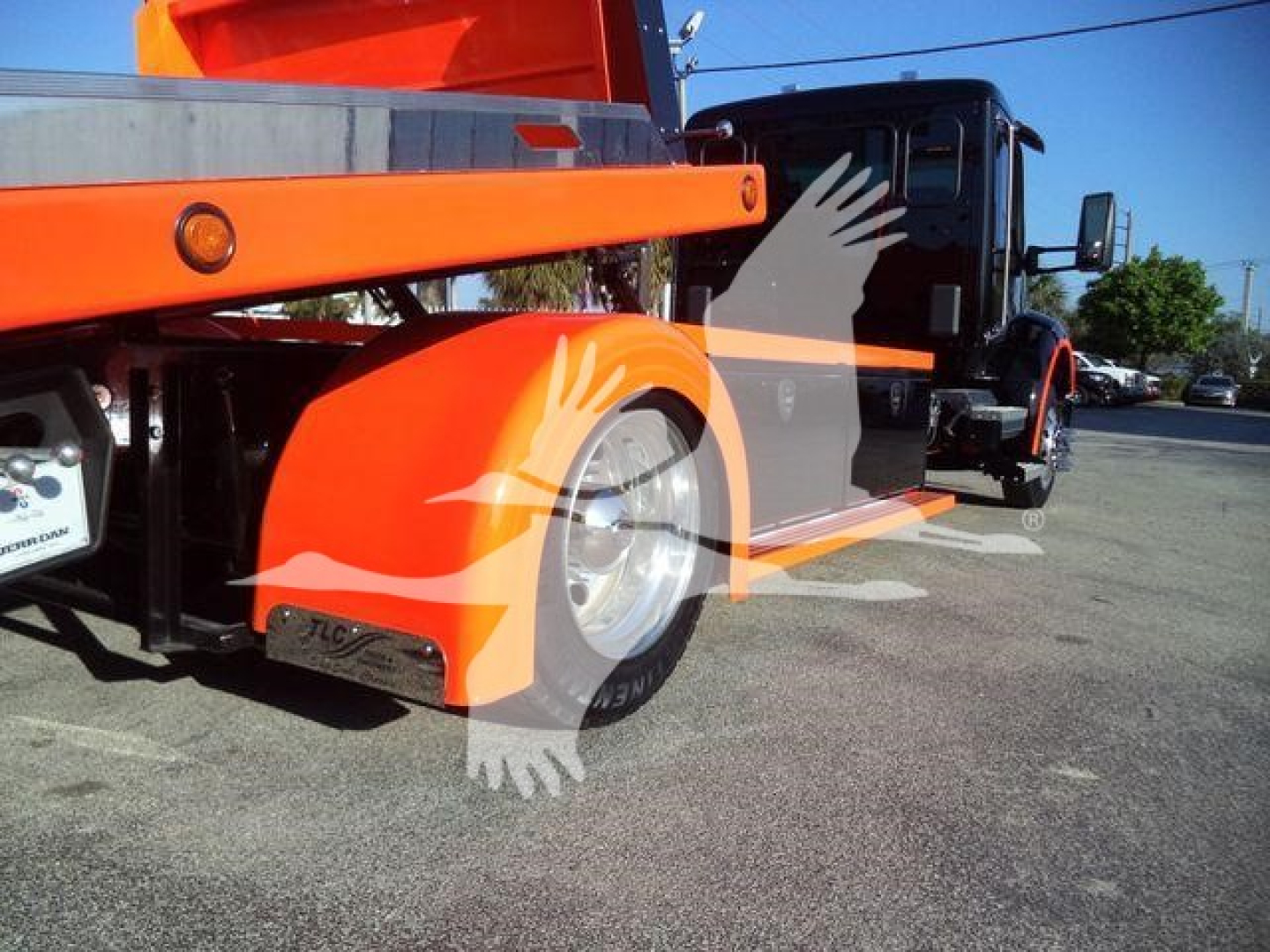 Kenworth T280 rollback tow truck in orange and black, side view showing rear wheels and hydraulic bed; overlay watermark of a hand and bird across center.
