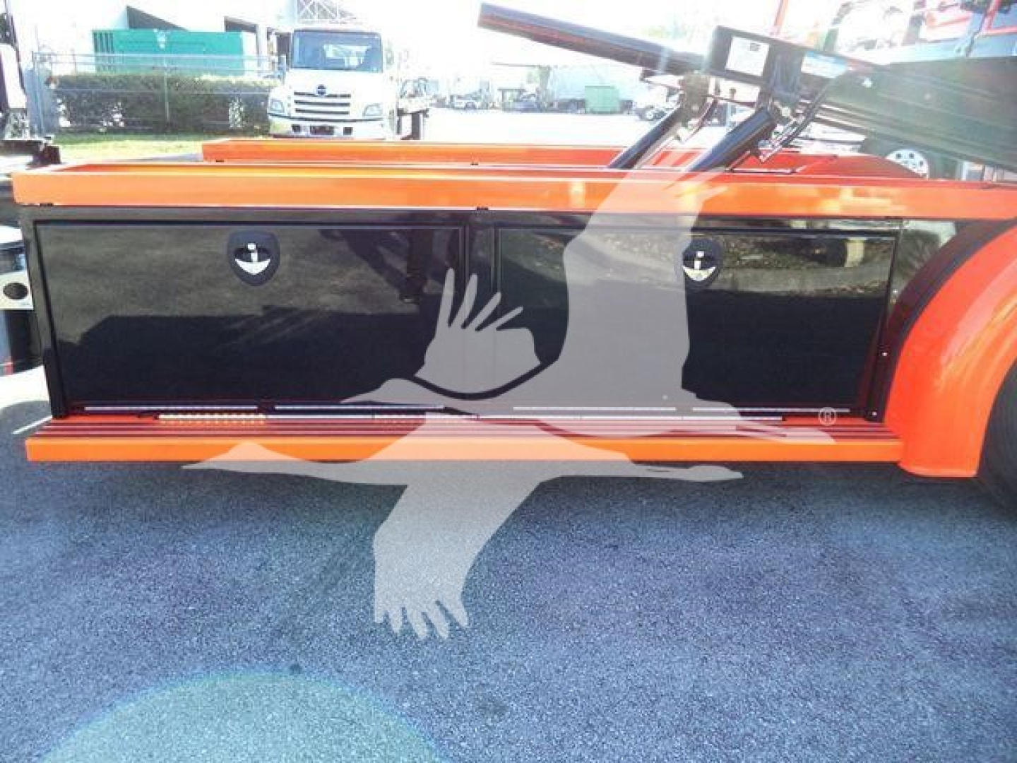 Kenworth truck side toolbox with glossy black panels and orange body. Two circular access doors on the panel. Reflection of a hand-shaped watermark overlay.