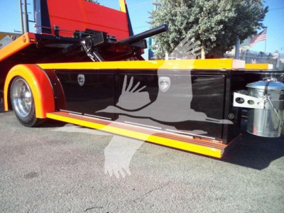 Kenworth T280 rollback truck side view showing black side panel with white graphic and yellow accents, storage box and chrome wheel; rear hitch and hydraulic components visible.