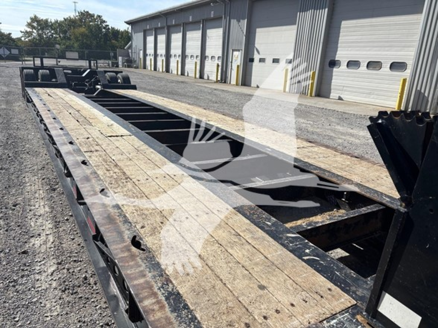 Lowboy trailer frame showing weathered wooden deck and steel frame on a gravel lot, with storage buildings in background.