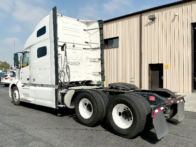 Truck tractor chassis and cab in a warehouse lot, showing rear sleeper area, tandem rear axles, and mudflaps.