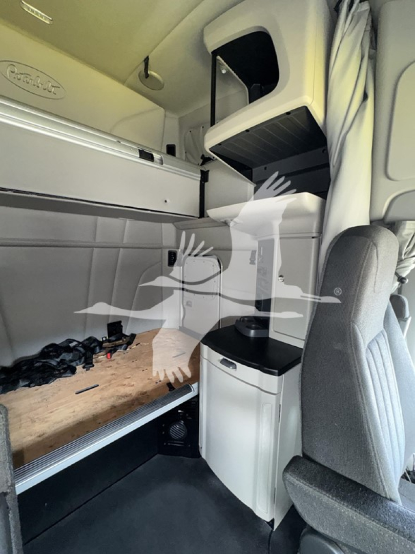 Peterbilt truck cab interior showing sleeper berth and storage compartments, with gray upholstered seat, overhead storage, and a small countertop cabinet near the bunk.