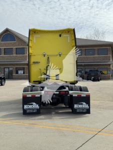 Rear view of a yellow Peterbilt 579 sleeper cab truck without trailer, showing back of cab and chassis with mud flaps and dealership stickers.