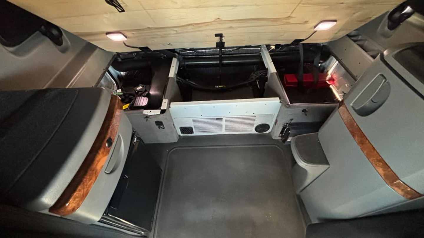 Truck cab interior showing sleeper area with storage compartments and panels, driver side curved wood trim, grey upholstery, and overhead lights.