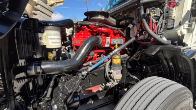 Diesel truck engine bay showing a red engine block with black hoses and yellow cap; visible labels include power steering reservoir and other components; front wheel well and radiator area.