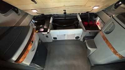 Truck cab interior showing sleeper area with storage compartments and panels, driver side curved wood trim, grey upholstery, and overhead lights.