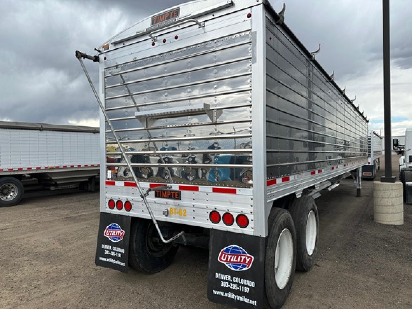 Trailer rear view showing 2024 Timpte Super Hopper with metal siderails and dual rear axles, utility decals and license plate area.