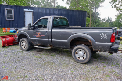 Ford F-250 Super Duty pickup with plow, 4x4 badge, gray cab and utility side decals, long bed, on gravel with blue building in background