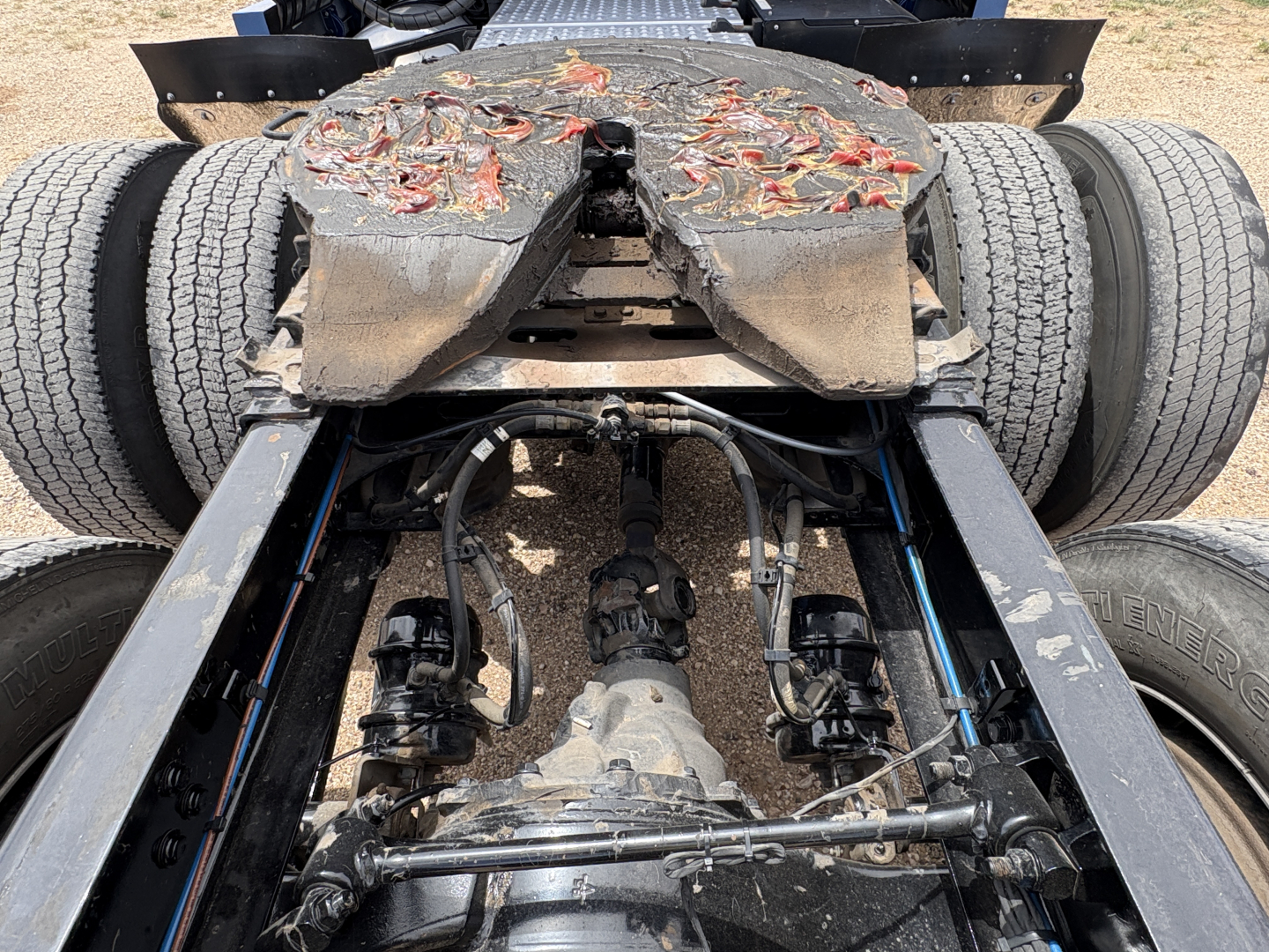 Tractor-trailer chassis with fifth-wheel and drivetrain components visible; dual rear wheels, suspension, and driveshaft; mud and wear on platform.