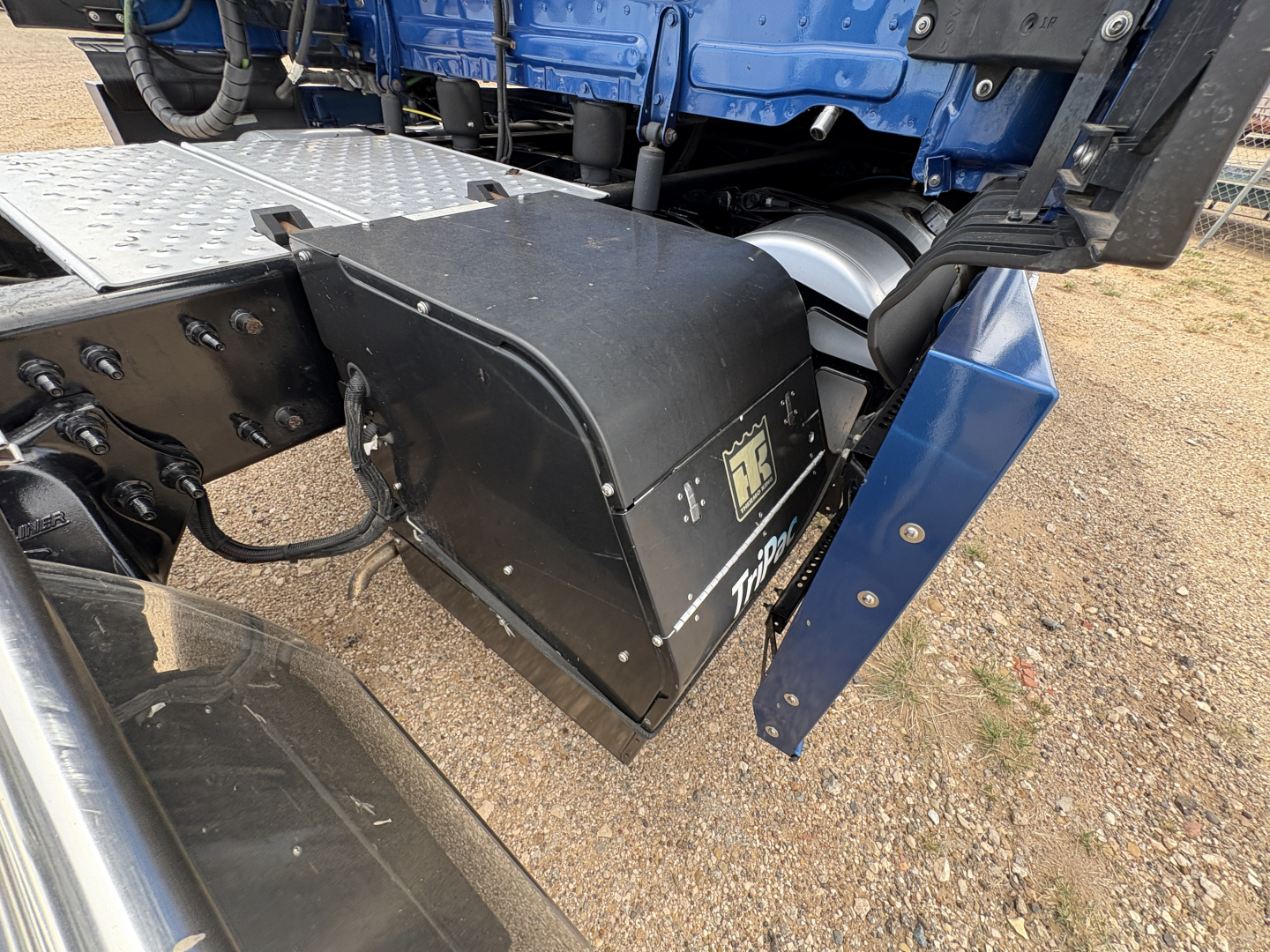 Truck component bay showing undercarriage area with black storage box and blue frame, Freightliner CASCADIA 126 cab seen, wiring and metal steps nearby.