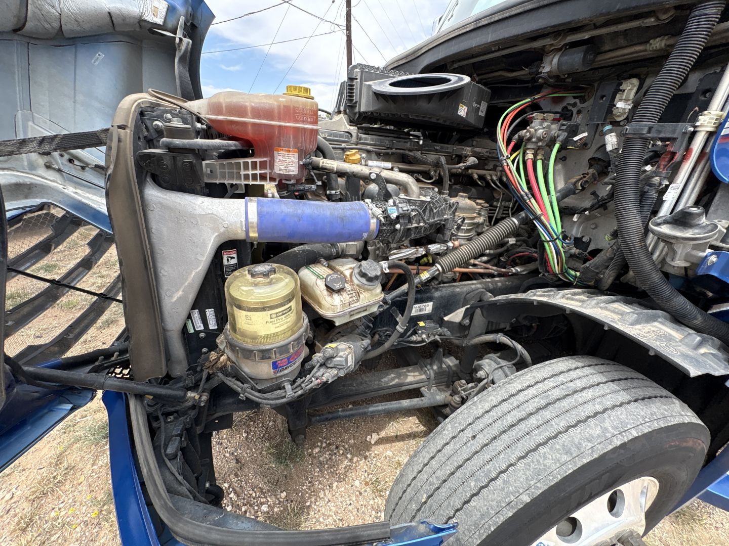 Truck engine bay of a 2022 Freightliner Cascadia 126 showing front left engine components, hoses, wiring harness, coolant reservoir, and air intake area.