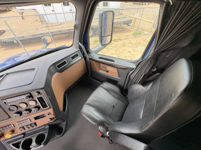 Dashboard and driver seat area of a 2022 Freightliner Cascadia 126 sleeper truck interior with black vinyl seats, beige dash panel, and brown trim, visible window and side mirror.