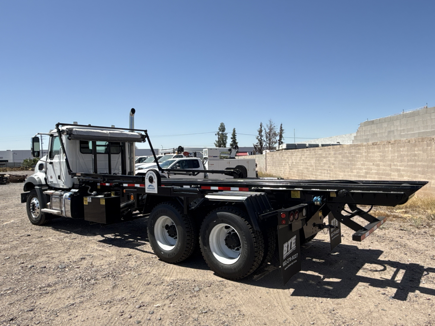 Mack GR64B roll-off truck shown in side view with long flatbed and dual rear axles, cab and chrome exhaust, black equipment, and clear blue sky.