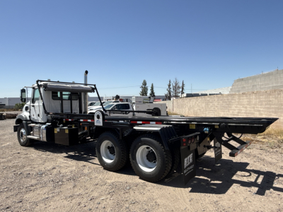 Mack GR64B roll-off truck shown in side view with long flatbed and dual rear axles, cab and chrome exhaust, black equipment, and clear blue sky.