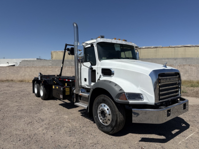 Truck: 2026 Mack GR64B rolloff in white, front-right view showing large chrome bumper, grille, tall exhaust stack, and flatbed/dolly setup.