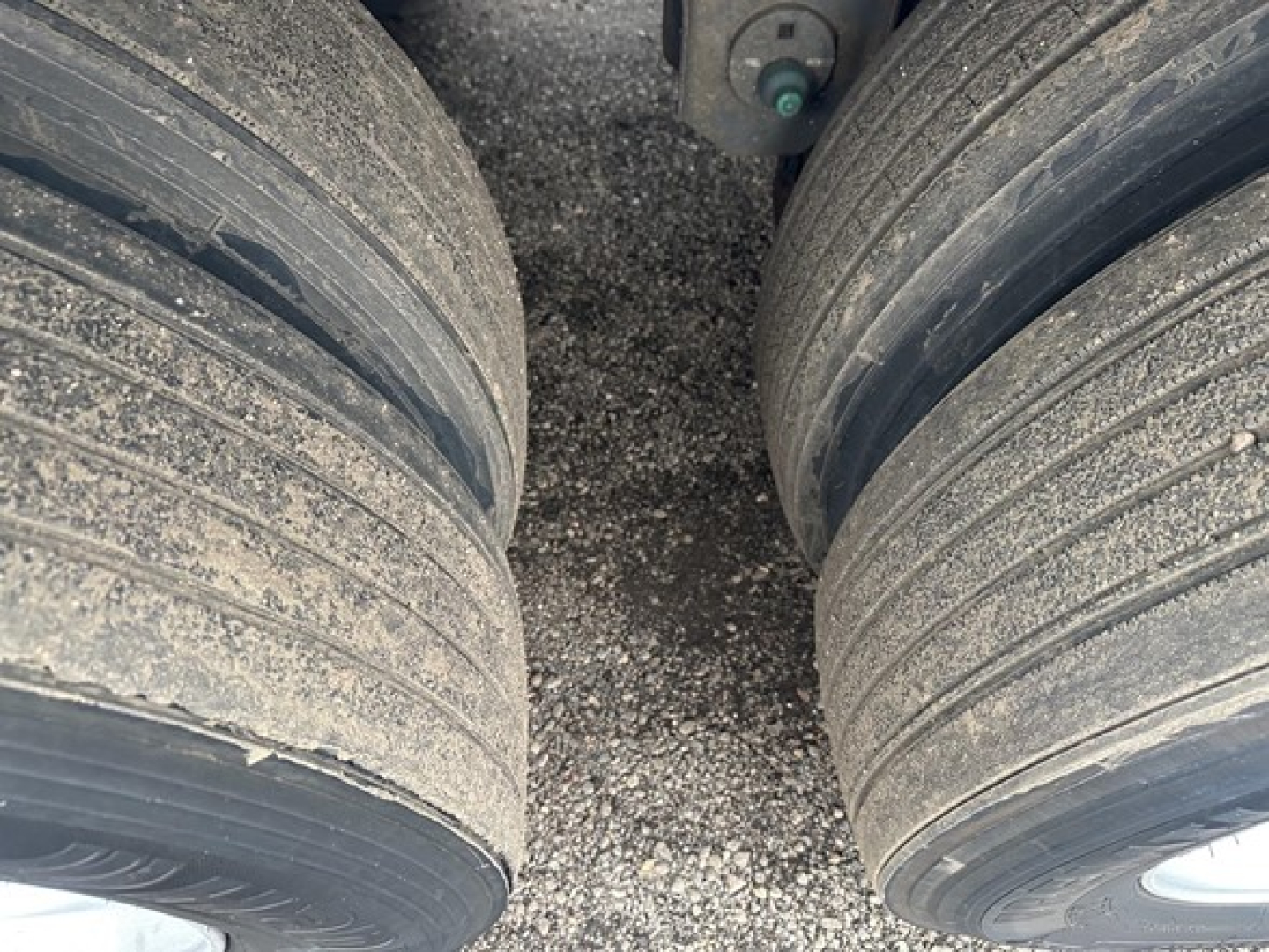 Trailer dual axle tires with rims and leaf spring undercarriage, gravel ground, close view of tires and undercarriage components.