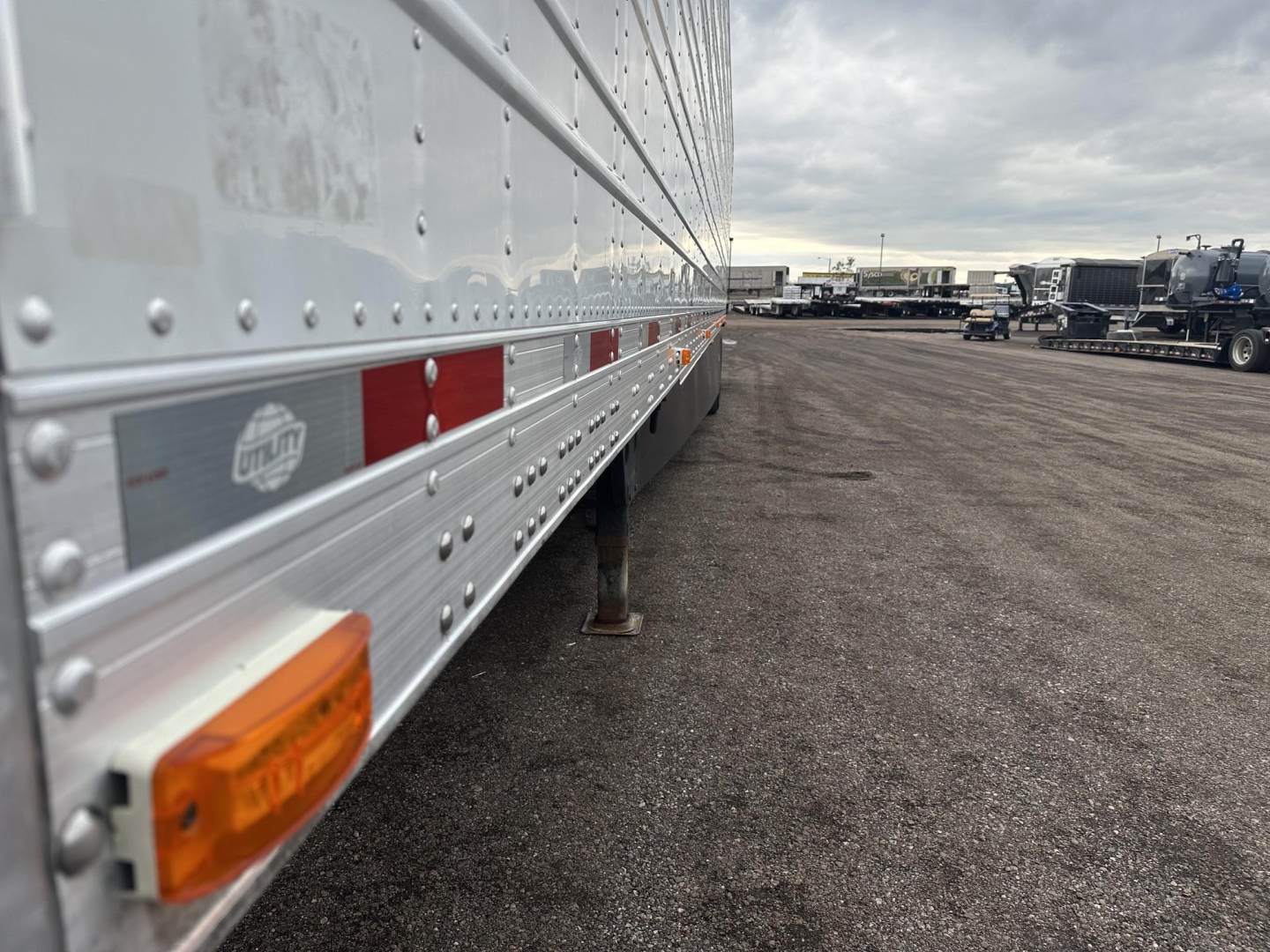 Side view of a 2023 Utility 3000R trailer showing lower side rails, reflective strips, riveted aluminum wall, and orange side marker light; distant lot with other trailers.