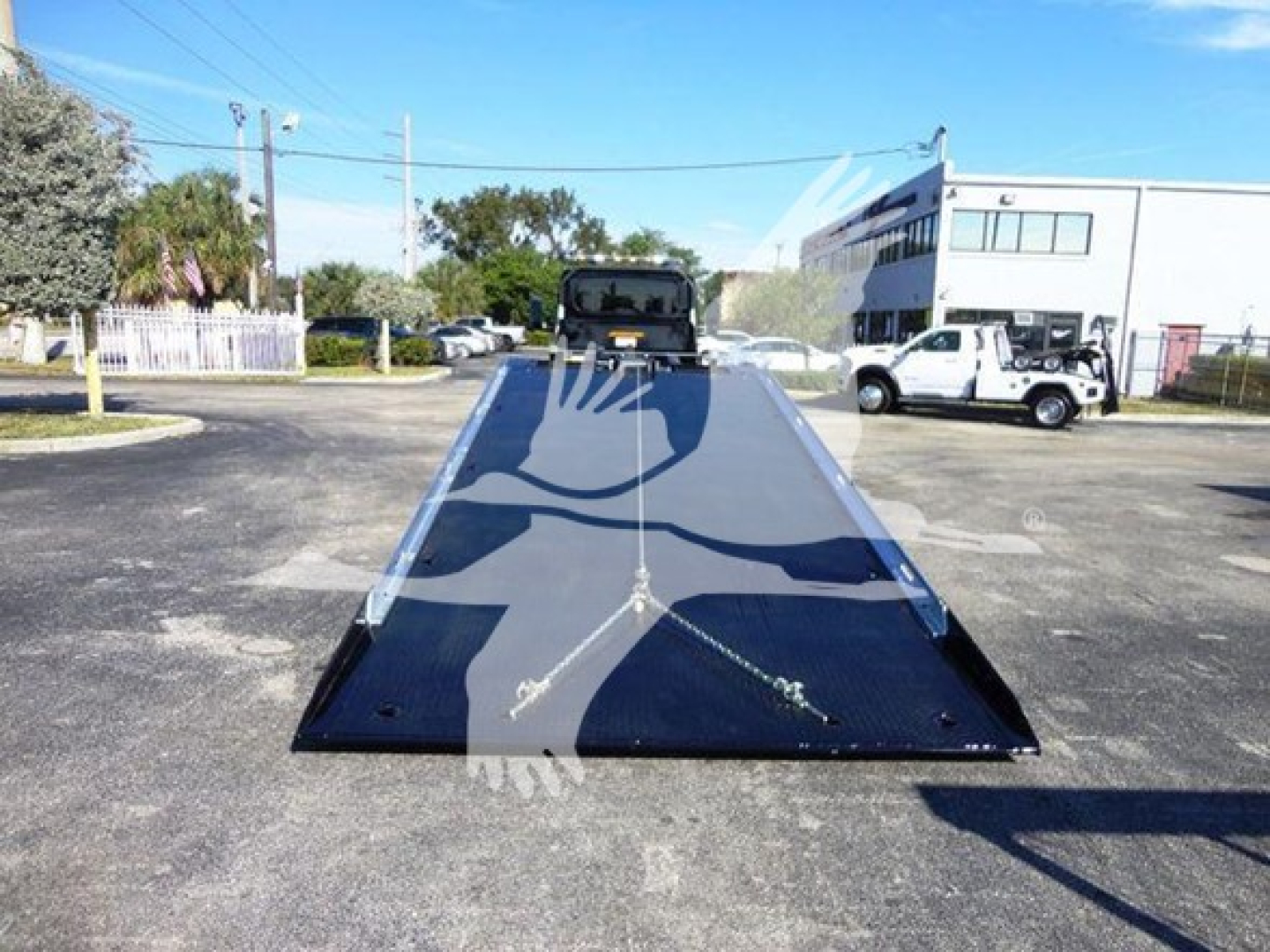 Flatbed tow truck with a long black loading surface and raised gooseneck at the end; in the distance a white pickup and a building visible under clear blue sky.