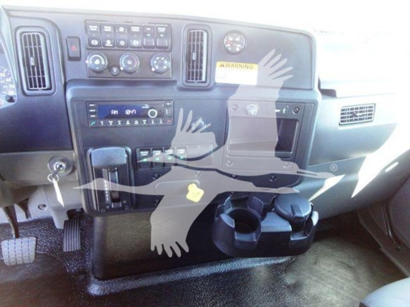 Truck cab dashboard with center console, knobs, vents, and cup holders; visible text includes control panel labels and a small warning sticker.