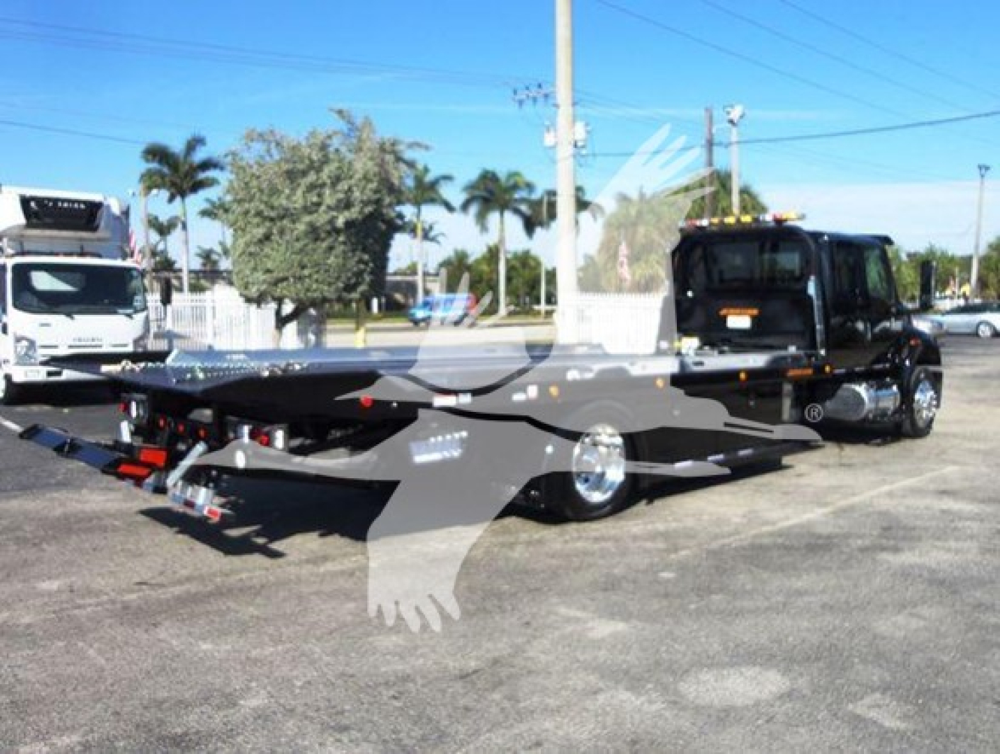 Flatbed rollback tow truck in a dealership lot with a black cab and gray bed, stock number J15011, International MV vehicle visible.