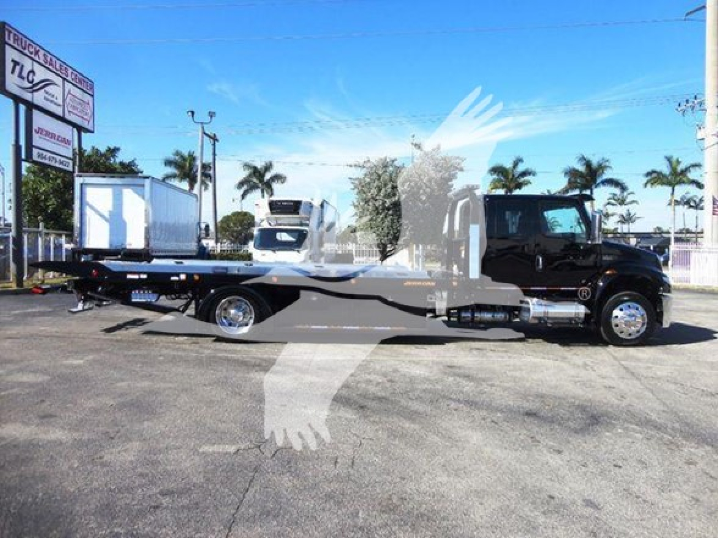 Flatbed tow truck in a dealership lot with palm trees in the background and a blue sky.