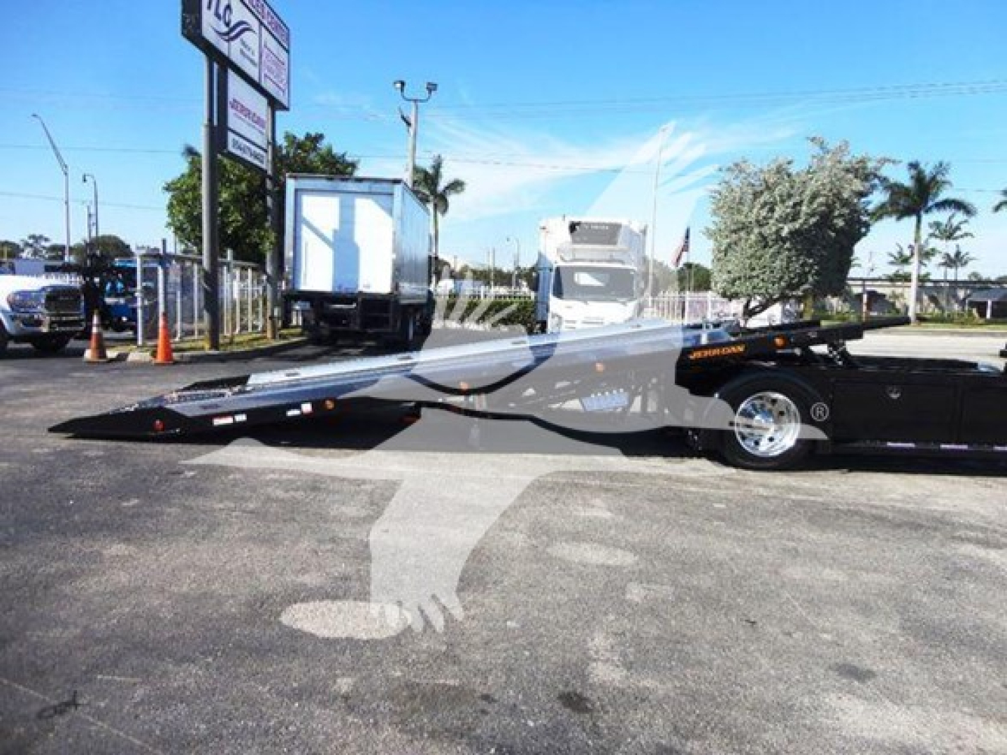 Truck rollback trailer in a dealership lot with a black flatbed, cab of a semi-truck in background and palm trees.