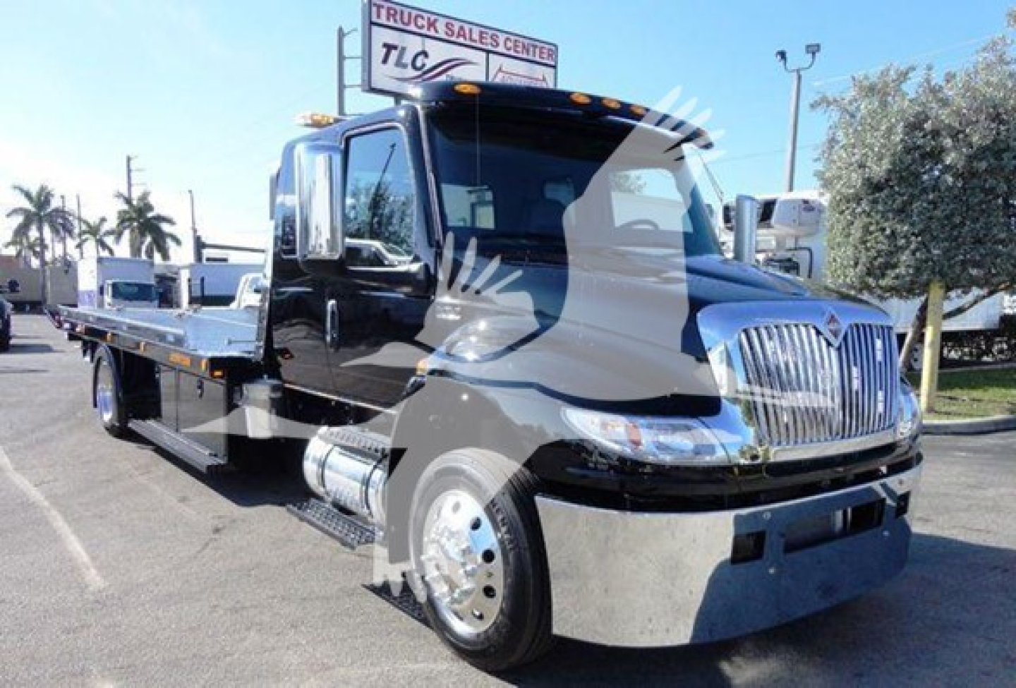 Black International MV rollback tow truck with extended flatbed, chrome grille, large side mirrors, and dual rear wheels; dealership signage is faint in background.