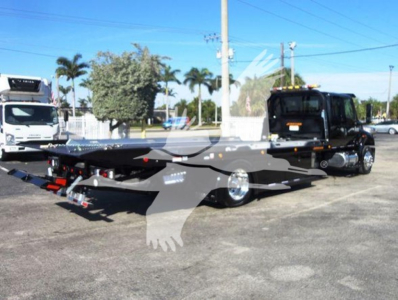 Flatbed rollback tow truck in a dealership lot with a black cab and gray bed, stock number J15011, International MV vehicle visible.