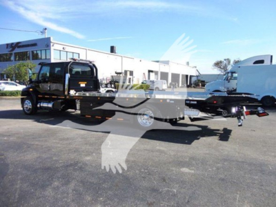 Truck bed rollback tow truck attached to a black heavy-duty tow vehicle with a long trailer, dealership lot in background.