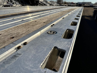 Flatbed trailer bed with aluminum side with multiple recessed tie-down slots and wooden deck visible from edge perspective.
