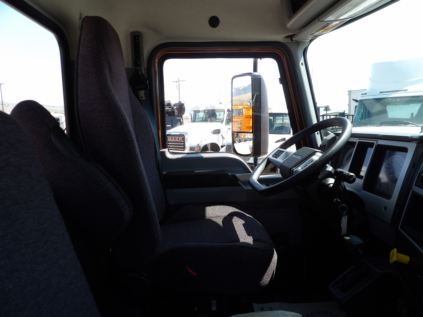 Cab interior of a 2026 Mack MD7 dump truck showing driver seat, steering wheel, dashboard, and side window with other trucks visible outside.