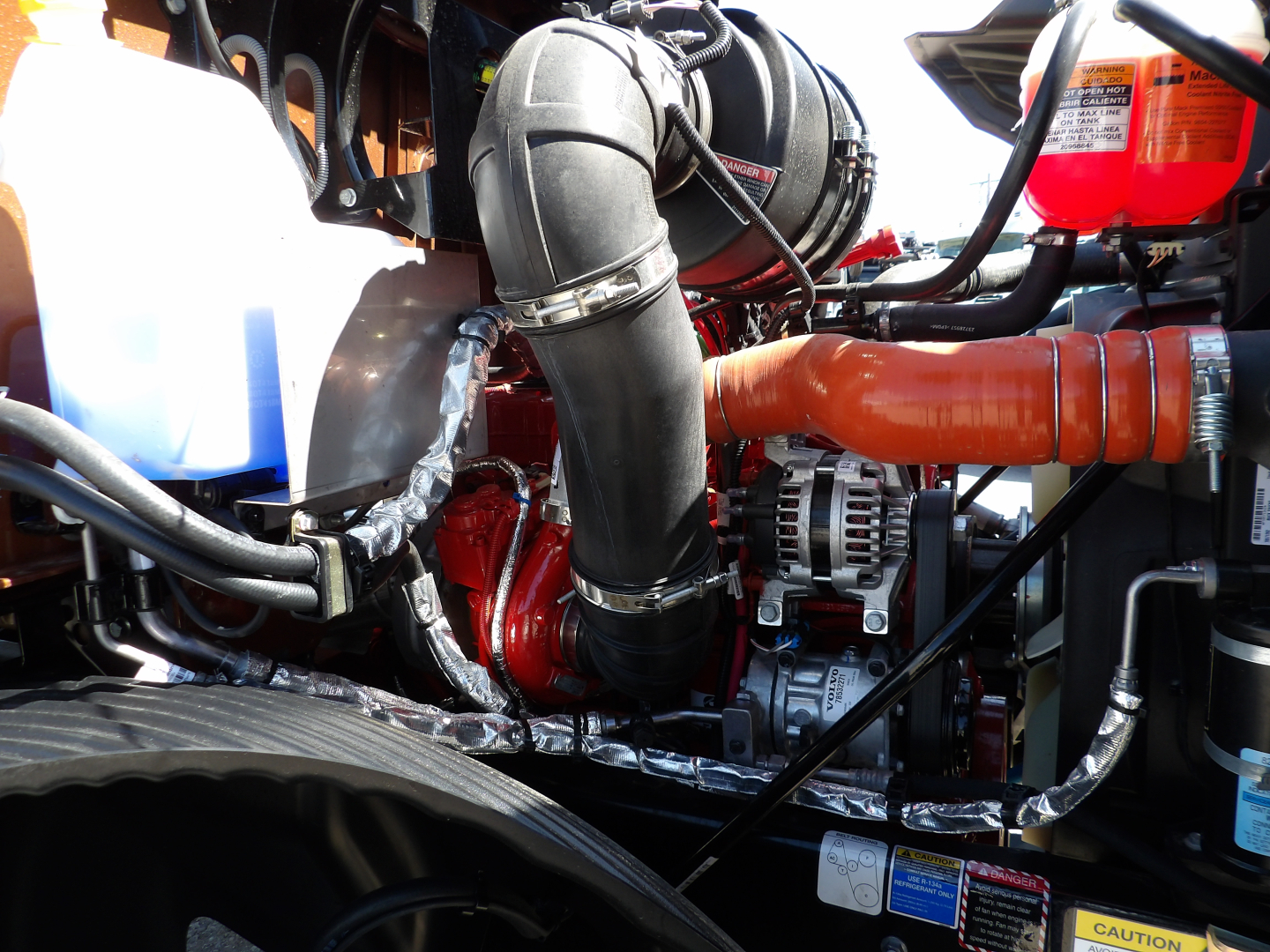 Vehicle engine bay of a 2026 Mack MD7 dump truck showing large black intake hose, orange exhaust/heat shield, alternator, coolant reservoir, and various hoses and wiring.